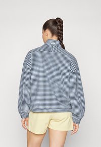 Ελεγμένο, oversized μπουφάν σε ναυτικό μπλε και λευκό, με ψηλό γιακά και ελαστικούς καρπούς, συνδυασμένο με κίτρινες βερμούδες. Διαθέτει χαρακτηριστικό λογότυπο.