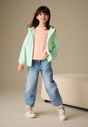 Jeune fille debout avec les mains dans un sweat à capuche zippé vert menthe, t-shirt couleur pêche, jean bleu ample et baskets à motif floral sur fond neutre.