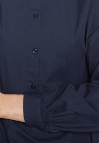 Camisa azul marinha de botões com gola, apresentando seis botões pretos e detalhes de punhos franzidos nas mangas longas. Textura de tecido suave.