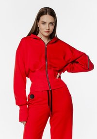Czerwona bluza z kapturem na zamek, z wysokim kołnierzem, długimi rękawami i ściągaczami, w zestawie z pasującymi czerwonymi spodniami z czarnym akcentem.