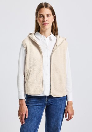 Beige fleece-liivi, jossa on huppu, täyspitkä vetoketju, sivutaskut ja teksturoitu, pehmeä kangas, joka on puettu valkoisen paidan päälle.