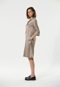 Braun gemustertes Kleid mit Kragen, 3/4-Ärmeln und Seiten Taschen. Das geometrische Design zeigt helle und dunkle Farben und ist aus einem glatten Stoff gefertigt.