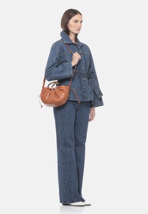 Donna che indossa una giacca di jeans con cintura e jeans, che tiene una borsa a tracolla in pelle marrone con manici rotondi.