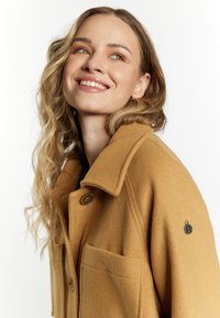 Cappotto in misto lana color cammello con collo a pistagna, due tasche frontali e una toppa decorativa sulla manica. Texture morbida e vestibilità su misura.