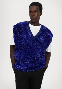 Mėlynas pūkuotas liemenė su V formos iškirpte, pagaminta iš tekstūruotos medžiagos, nešiojama ant baltos marškinėlių, derinama su juodomis kelnėmis.