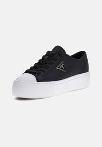 Zapatilla baja negra con suela de plataforma gruesa blanca, puntera blanca, cordones negros y logo triangular de metal en el lateral.