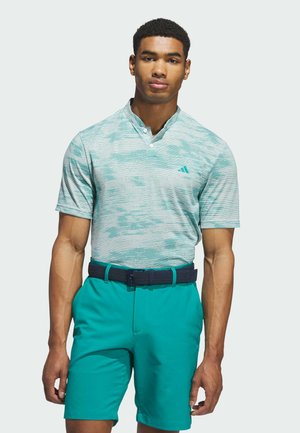 Man wearing teal patterned short-sleeve polo shirt and teal shorts with navy woven belt, standing against plain light background.