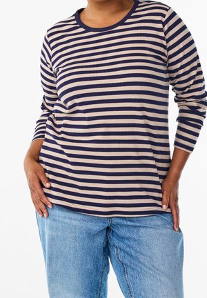 Personne portant un haut à manches longues rayé horizontalement beige et bleu marine et un jean en denim bleu clair, les mains reposant sur les hanches.