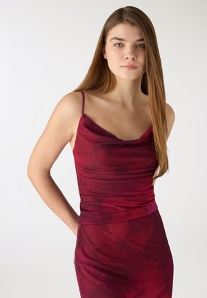 Giovane donna con capelli lunghi e lisci, che indossa un vestito senza maniche color bordeaux con spalline sottili, in piedi contro uno sfondo chiaro e semplice.