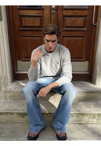 Fiatal férfi szürke pulóverben és kék farmerral, betonjárdán ül egy faajtó előtt, lefelé nézve, gondterhelt arckifejezéssel.