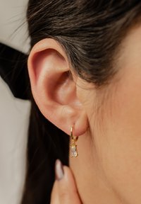 Orecchino a cerchio dorato con una pietra rettangolare trasparente. L'orecchino pende dal lobo dell'orecchio, mettendo in risalto una finitura in metallo lucido e un design semplice.