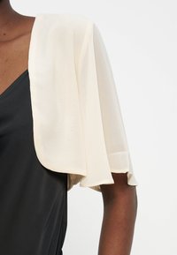 Κοντινό πλάνο ενός ατόμου που φοράει μαύρο τοπ χωρίς μανίκια με μια διάφανη, εκρού κάπα κρεμασμένη πάνω στον έναν ώμο και το πάνω μέρος του χεριού.