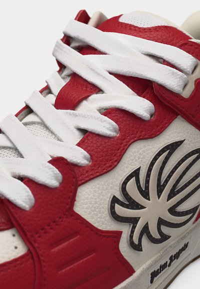 Κοντινό πλάνο κόκκινου και λευκού αθλητικού παπουτσιού με λευκά κορδόνια και μαύρο στυλιζαρισμένο λογότυπο αράχνης στην υφασμένη δερμάτινη πλαϊνή επιφάνεια.