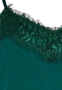 Satin-top i smaragdgrön färg med intrikata spetsdetaljer med blommönster och fransar längs halsringningen. Slät textur.