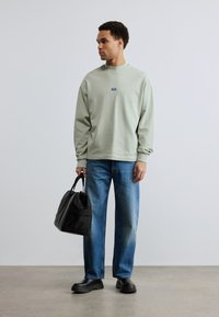 Νεαρός άνδρας με ανοιχτό πράσινο φούτερ και μπλε τζιν κρατά μια μαύρη τσάντα, στέκεται σε απλό πάτωμα μπροστά σε γκρι τοίχο.