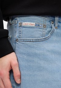 Heledad sinine teksapüksid, millel on sile tekstuur, punase ja valge "Calvin Klein" silt vöökohal ning hõbedased riistvara aktsendid.