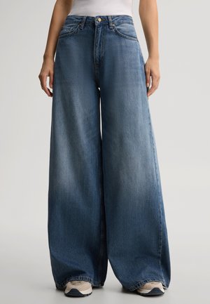 Persoon draagt blauwe jeans met wijde pijpen en witte sneakers, staand tegen een effen achtergrond.