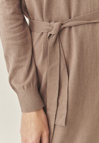 Savunruskea neulepaita, jossa on solmittava vyö vyötäröllä, resorireunaiset hihansuut ja pehmeä tekstuuri, korostaen yhtenäistä, viihtyisää muotoilua.