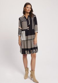 Czarna i beżowa sukienka w paski z kołnierzykiem zapinanym na guziki, rękawami długości trzy czwarte oraz falbanowym dołem. Para beżowych botków na obcasie.