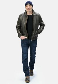 Braune Lederjacke mit hohem Kragen, schwarzes Hemd, blaue Jeans und braune Stiefel. Der Mann trägt außerdem eine blaue Strickmütze.