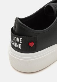 Μαύρο αθλητικό παπούτσι με υφή δερμάτινης εξωτερικής επιφάνειας, λευκή καουτσούκ σόλα, επώνυμο ύφασμα και μια μικρή κόκκινη καρδιά ως διακόσμηση.