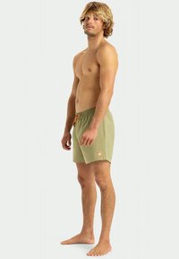 Πράσινα μαγιό με κορδόνι στη μέση, με λεπτή υφή και μια μικρή ετικέτα λογότυπου στην αριστερή πλευρά. Ορατά τα γυμνά πόδια του μοντέλου.