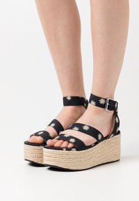Voeten in zwarte plateau-wedge-sandalen met madeliefjespatronen en enkelriemen op een witte achtergrond.