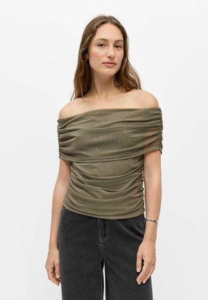 Jeune femme portant un haut froncé vert olive épaules dénudées et un jean noir, debout devant un fond blanc uni.