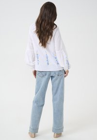 Blusa di lino bianca con ricami floreali blu e gialli, maniche a sbuffo, abbinata a jeans a gamba larga di un blu chiaro e scarpe beige.