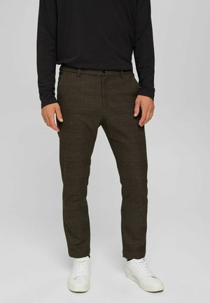Mann trägt braune karierte Slim-Fit-Hose, schwarzes Langarmshirt und weiße Sneakers und steht vor einem einfarbigen Hintergrund.