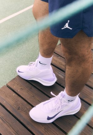 Chaussures de sport blanches et lavande avec une tige texturée, dotées d'un swoosh Nike violet et d'un système de laçage, portées avec des chaussettes blanches sur un sol en bois.