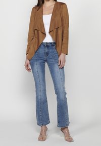 Femme portant une veste marron drapée sur un haut blanc, un jean bleu taille haute à jambes droites et des sandales à talons translucides.