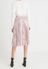 Rosa plissierter Midirock mit glänzender Textur und elastischem Taillenbund. Kombiniert mit einem weißen Langarmshirt und schwarzen Sandalen mit Absatz.