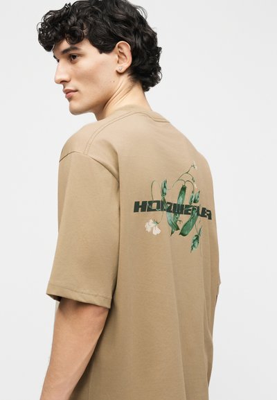 Νέος άνδρας με σκούρα, σγουρά μαλλιά, που φοράει μπεζ μπλουζάκι με πράσινο γραφικό φυτού μπιζελιού και μαύρο κείμενο στο πίσω μέρος, κοιτάζοντας πλάγια.