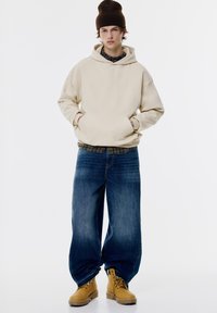 Beige hoodie med en framficka, bärs över en mörk flanellskjorta; stora blå jeans och gula arbetsskor fulländar looken.