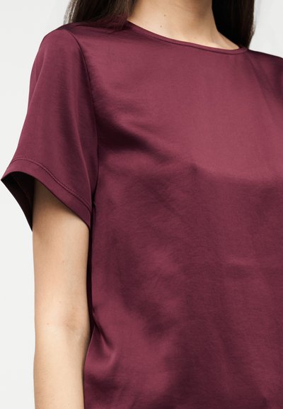 Μπορντό μπλούζα με κοντά μανίκια και στρογγυλή λαιμόκοψη, με λεία υφή και διακριτική λάμψη. Διαθέτει ευθείες, καθαρές ραφές στα μανίκια.