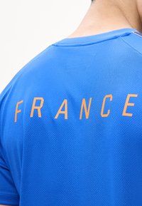 Άτομο που φορά μπλε πουκάμισο με υφασμάτινη υφή, πορτοκαλί κείμενο "FRANCE" στο πάνω μέρος της πλάτης, ορατός λαιμός και μερική πλευρά του κεφαλιού.