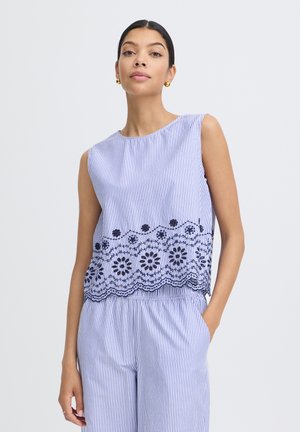 Femme portant un haut sans manches à rayures verticales bleues et blanches avec une broderie florale noire, accompagné d'un pantalon ample assorti, debout avec une main dans la poche.