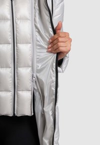 Ezüst színű, steppelt mintázatú pufi dzseki, dupla cipzárral és sima, fényes anyagból; belső bélése gyermekesen fekete.