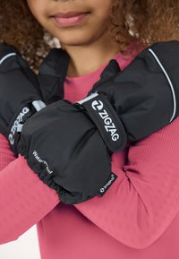 Sorte vandtætte handsker med et zigzag-logo og reflekterende detaljer, båret over en tekstureret pink langærmet skjorte. Nærbillede af hænder, der krydser hinanden.