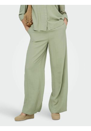 Ember világoszöld, bő szárú nadrágban és hozzá illő, gombos ingben, zsebében tartja a kezét, és egy fonott táskát fog.