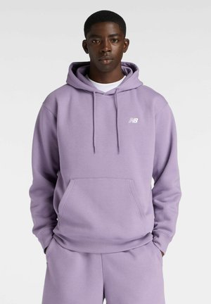 Junger Mann mit dunkler Haut, der einen hellvioletten New Balance Hoodie und passende Jogginghose trägt, steht mit den Händen in den Taschen.