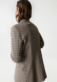 Blazer à carreaux marron et beige avec une coupe ajustée, des revers crantés et une poche unique. Tissu texturé avec des motifs à carreaux contrastants.