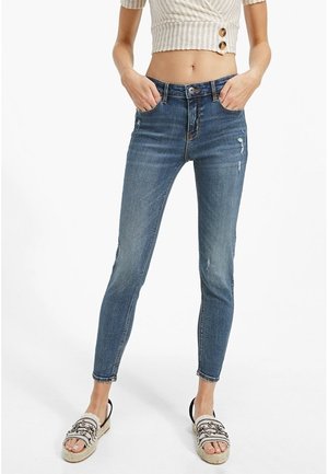 Vrouw draagt een high-waist skinny blauwe spijkerbroek met versleten details, een cropped gestreept topje met knopen, en gevlochten sandalen met kraalversieringen.