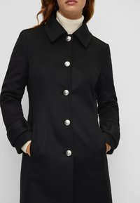 Cappotto di lana nera con scollo a collare, bottoni argentati e tasche laterali. Presenta una texture liscia e una silhouette dritta.