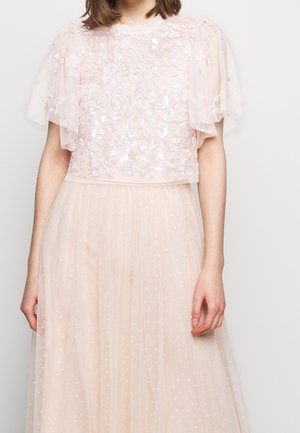 Frau trägt ein hellrosafarbenes Spitzenkleid mit floralen Pailletten und transparenten, kurzen Flatterärmeln, steht vor einem schlichten hellen Hintergrund.