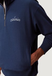 Sudadera azul marino con cuello de cremallera de cuarto, logo "calvin" bordado, bolsillos laterales y puños acanalados para un ajuste seguro y comodidad.