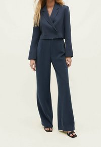 Tumma laivastonsininen kaksiosainen puku, johon kuuluu lyhyt kaksirivinen bleiseri leveillä hihoilla ja korkeavyötäröiset leveälahkeiset housut, valmistettu sileästä kankaasta.