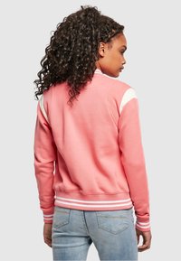 Ružičasta majica dugih rukava s bijelim naglascima na ramenima, rebrastim prugastim manžetama i rubom. Mekani materijal i uski kroj. Prikazan je pogled s leđa.