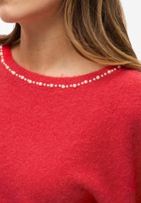 Maglione rosso in lana con scollo rotondo decorato da una fila di piccole perle finte bianche. Il tessuto morbido e texturizzato è visibile.